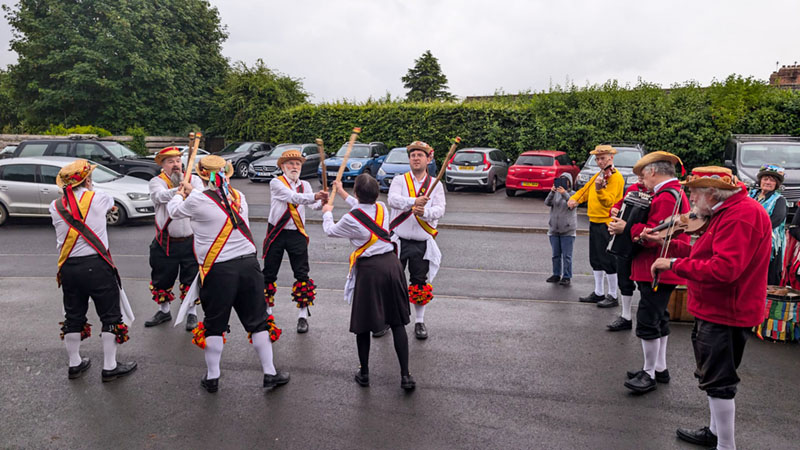 6 Shakespeare MOrris Dancers in their Cotswald attire and sticks dancing in front of a row of parked cars