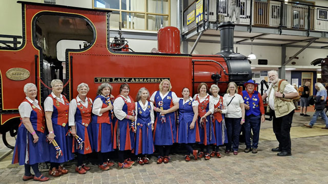 dancers and musicians lined up in front of a bright red steam engine in the sheds at Highley