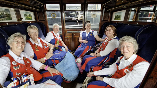 6 dancers sitting in a first class compartment, luxuriously fitted out with rich coloured fabric and pictures in frames